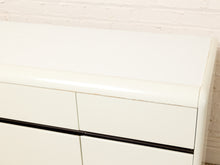 Load image into Gallery viewer, Image 13 of 18: Close-up of a glossy white dresser or cabinet with a curved top edge and a black horizontal handle, set against a white brick wall.