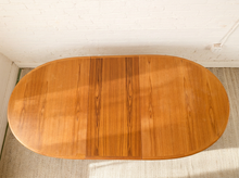 Load image into Gallery viewer, Image 9 of 28: An oval wooden table with a warm honey-toned finish and visible wood grain, photographed from above against a white brick wall and light rug.