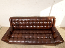 Load image into Gallery viewer, Image 4 of 13: A dark brown tufted leather loveseat sofa with a high back, shown against a white brick wall and light-colored floor.