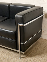 Load image into Gallery viewer, Image 5 of 14: A close-up of a black leather Chesterfield-style sofa with a polished chrome tubular steel frame, shown on a textured beige carpet against a white brick wall.