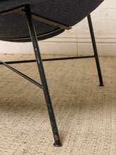 Load image into Gallery viewer, Image 16 of 17: Close-up of a mid-century modern chair with a dark textured fabric seat and black metal legs, set on a beige woven rug against a white brick wall.