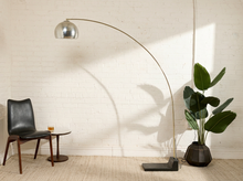 Load image into Gallery viewer, Image 7 of 23: A modern arched floor lamp with a brushed silver dome shade and a dark rectangular base stands in a bright room with a white brick wall. A black leather chair, a small round side table, and a potted green plant are arranged around it.
