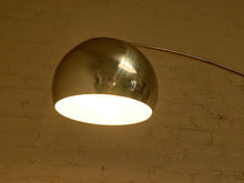 Load image into Gallery viewer, Image 10 of 23: A polished metallic dome floor lamp with a curved arm, shown illuminated against a warm-toned brick wall background.