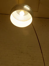 Load image into Gallery viewer, Image 14 of 23: A modern floor lamp with a curved dark metal stem and a spherical shade, glowing with a warm yellow light against a beige brick wall.