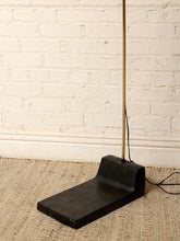 Load image into Gallery viewer, Image 19 of 23: A modern black concrete floor lamp with a wide rectangular base and a slim vertical brass-colored pole, standing on a woven jute rug against a white brick wall.