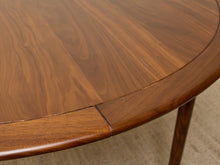 Load image into Gallery viewer, Image 7 of 12: Close-up of a round wooden table with a warm medium-brown finish, showing the wood grain and a seam near the curved edge, with part of a table leg visible on a light neutral floor.
