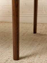 Load image into Gallery viewer, Image 12 of 12: Close-up of a dark brown wooden table leg standing on a woven beige rug in front of a white brick wall.