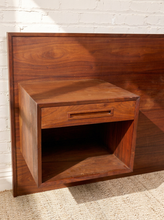 Load image into Gallery viewer, Image 7 of 22: A mid-century modern walnut nightstand with a single drawer and open shelf, set against a white brick wall and light beige carpet.