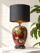 Load image into Gallery viewer, Image 1 of 19: Vintage 1950s table lamp featuring a rounded burgundy base with hand-painted yellow and cream flowers, green foliage, and decorative bird motifs, resting on a dark pedestal base and topped with a black cylindrical fabric shade.