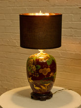 Load image into Gallery viewer, Image 4 of 19: A decorative table lamp with a dark brown fabric drum shade and a glossy dark red ceramic base decorated with gold, green, and yellow floral and bird motifs, shown lit against a white brick wall.