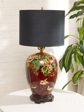 Load image into Gallery viewer, Image 5 of 19: A decorative table lamp with a glossy dark red ceramic base decorated with gold cranes and floral motifs, topped with a black fabric drum shade and a dark wooden footed stand.