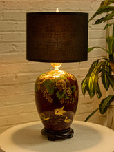 Load image into Gallery viewer, Image 6 of 19: A decorative table lamp with a dark brown ceramic body featuring hand-painted floral and crane motifs, topped with a black fabric drum shade and resting on a dark wooden base.