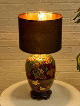 Load image into Gallery viewer, Image 8 of 19: A decorative table lamp with a dark brown fabric drum shade and a glossy ceramic base featuring a floral and bird pattern in gold, green, and brown, set against a white brick wall.