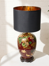 Load image into Gallery viewer, Image 7 of 19: A decorative table lamp with a black fabric drum shade, a glossy dark red ceramic vase body decorated with gold floral and bird motifs, and a dark wooden base, shown on a white pedestal against a white brick wall.