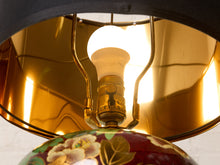 Load image into Gallery viewer, Image 12 of 19: Close-up of a decorative table lamp with a glowing white bulb, a polished gold metal shade, and a dark red floral ceramic base.