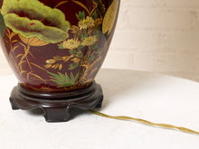 Load image into Gallery viewer, Image 19 of 19: A close-up of a decorative ceramic lamp base with a glossy dark red body, hand-painted lotus and floral motifs in gold, green, and yellow, mounted on a dark wooden stand.