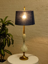 Load image into Gallery viewer, Image 4 of 19: A table lamp with a dark blue-gray textured drum shade, a gold-finished stem and base, and a glossy white sculptural bulb-shaped center, shown lit on a round white table against a beige brick wall with a green plant at the side.