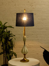 Load image into Gallery viewer, Image 8 of 19: A table lamp with a dark navy blue textured drum shade, a gold-finished stem and base, and a cream-colored ribbed bulb-shaped ceramic body, shown lit on a white round table against a white brick wall.