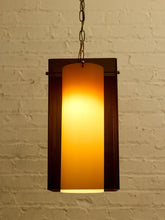 Load image into Gallery viewer, Image 4 of 24: A rectangular hanging pendant light with a frosted warm yellow glass shade and dark metal frame, suspended by a chain against a white brick wall.