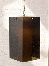 Load image into Gallery viewer, Image 9 of 24: A modern rectangular pendant light with a translucent dark amber-brown smoked glass shade and a brass chain, hanging against a white brick wall.
