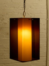 Load image into Gallery viewer, Image 10 of 24: A hanging pendant light with a dark amber-brown translucent outer shade and a glowing warm yellow-orange inner shade, suspended against a white painted brick wall.
