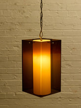Load image into Gallery viewer, Image 2 of 24: A hanging rectangular pendant light with a glowing warm yellow-orange cylindrical shade inside a dark amber-brown translucent frame, suspended by a chain against a white brick wall.