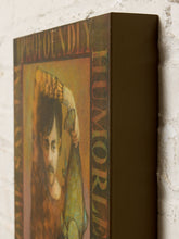 Load image into Gallery viewer, Image 6 of 9: A close-up of a framed or mounted art print leaning against a white brick wall. The artwork features a portrait with the words 'PROFOUNDLY HUMOROUS' visible along the top and side.