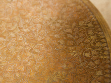 Load image into Gallery viewer, Image 7 of 13: Vintage round brass-tone foot warmer featuring a circular top with intricate floral and scrollwork engravings, a raised rounded rim, warm aged patina, and three short turned metal legs.