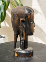 Load image into Gallery viewer, Image 7 of 13: A dark carved wooden African-style head sculpture with a high ridged back and long ear, displayed on a round black table against a light wall with a plant leaf in the background.