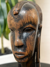 Load image into Gallery viewer, Image 9 of 13: A close-up of a hand-carved wooden sculpture of a female face, showcasing rich dark brown and warm honey wood grain patterns, set against a softly blurred indoor background with a plant.