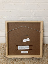 Load image into Gallery viewer, Image 9 of 9: The back of a framed art print showing a light wood frame, brown backing board, wire hanger, and informational labels, set against a white brick wall and woven rug.