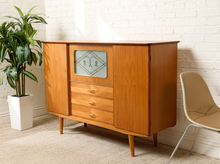 Load image into Gallery viewer, Image 2 of 27: A mid-century modern wooden sideboard cabinet with three drawers and a glass-front upper cabinet door, shown in a room setting against a white brick wall with a potted plant and a chair.