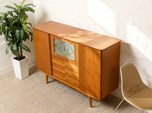 Load image into Gallery viewer, Image 3 of 27: A mid-century modern wooden sideboard cabinet with a warm honey finish, featuring three drawers with metal handles and a glass-front cabinet door with geometric patterns. The cabinet is styled in a room setting against a white brick wall, with a potted green plant on the left and a beige chair on the right.