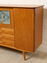 Load image into Gallery viewer, Image 6 of 27: Mid-century modern wooden sideboard cabinet with a warm honey-toned wood finish, featuring a glass-front cabinet door, three drawers with brass pulls, and tapered legs, shown against a white brick wall and textured carpet.