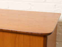 Load image into Gallery viewer, Image 9 of 27: Close-up of a mid-century modern wooden cabinet or side table showing the warm medium-brown wood grain and rounded corner of the top, set against a white brick wall background.