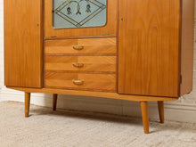 Load image into Gallery viewer, Image 16 of 27: Mid-century modern wooden cabinet with three drawers, two side doors, and a decorative glass panel at the top, shown on a carpeted floor against a white brick wall.