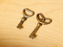 Load image into Gallery viewer, Image 18 of 27: Two vintage-style metal keys with heart-shaped bow loops resting on a light wood grain surface.