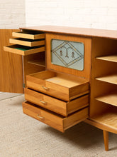Load image into Gallery viewer, Image 22 of 27: Mid-century modern wooden credenza or storage cabinet with multiple pull-out drawers, open shelving, and a decorative inset window panel, shown in a room setting with carpet and white brick wall visible.