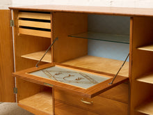 Load image into Gallery viewer, Image 24 of 27: A mid-century modern wooden writing desk or cabinet with a pull-out writing surface, glass shelf, and open storage compartments, shown against a white brick wall.