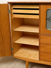 Load image into Gallery viewer, Image 27 of 27: A mid-century modern wooden cabinet with its left door open, showing light wood shelves and drawers on a beige carpeted floor.