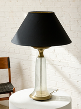 Load image into Gallery viewer, Image 3 of 18: A table lamp with a black fabric conical shade, a clear glass cylindrical base, and a brass-toned metal neck and base, displayed on a white round pedestal against a white brick wall.