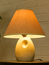 Load image into Gallery viewer, Image 6 of 14: A table lamp with a warm orange fabric shade and a cream-colored ceramic base featuring a circular cutout, shown lit against a white brick wall background.