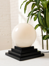 Load image into Gallery viewer, Image 5 of 22: A modern table lamp featuring a large, smooth, off-white spherical shade resting on a tiered black wooden base, set against a white brick wall with a green plant in the background.