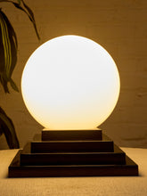 Load image into Gallery viewer, Image 8 of 22: A glowing spherical table lamp with a matte white globe and a dark tiered rectangular base, photographed indoors against a warm beige wall with a plant leaf visible at the left edge.
