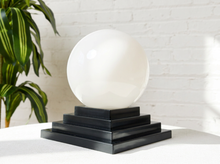 Load image into Gallery viewer, Image 11 of 22: A large smooth white spherical lamp or decorative orb displayed on a tiered black pedestal base, set on a white surface against a white brick wall with a green plant visible at the left edge.