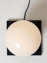 Load image into Gallery viewer, Image 17 of 22: A modern pendant light with a large, round, frosted off-white glass globe shade mounted on a square black metal base, shown from a top-down view against a light gray textured background.