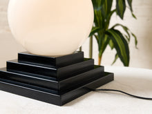 Load image into Gallery viewer, Image 20 of 22: A modern table lamp with a glossy black tiered square base and a large, smooth, spherical off-white shade, shown on a textured light surface with a blurred green plant in the background.