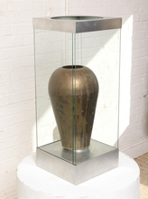 Load image into Gallery viewer, Image 4 of 18: Vintage dark brown sculptural vase with rounded shoulders, a narrow circular opening, tapered body, and weathered surface, displayed inside a tall rectangular glass case with brushed silver-tone metal top and base.