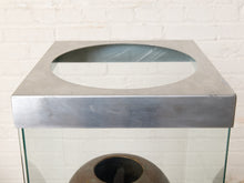 Load image into Gallery viewer, Image 7 of 18: Vintage dark brown sculptural vase with rounded shoulders, a narrow circular opening, tapered body, and weathered surface, displayed inside a tall rectangular glass case with brushed silver-tone metal top and base.