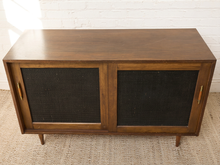 Load image into Gallery viewer, Image 8 of 24: Vintage wooden credenza with two black textured sliding cabinet doors, warm brown wood frame, gold-tone hardware, and short tapered legs
